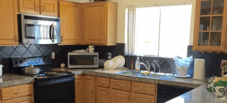 A kitchen with wooden cabinets, a black stove with a pot on it, a microwave above the stove, a countertop with a dish rack holding plates, a sink under a window with blinds, and various kitchen items including a dish soap bottle, paper towel roll, and a blue ice bucket.