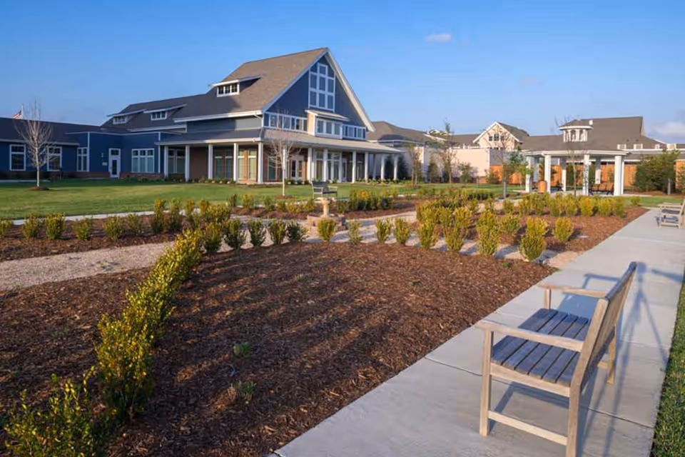 Outdoor view of The Crossings Retirement Community showing a landscaped garden with small bushes and mulch beds, a concrete walkway with wooden benches, and large residential-style buildings in the background under a clear blue sky.