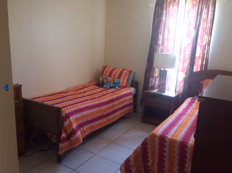 A small bedroom with two single beds covered in matching orange, pink, and white striped bedspreads. There are three stuffed animals on one bed. A wooden nightstand with a lamp is positioned between the beds, and a window with floral curtains lets in natural light.