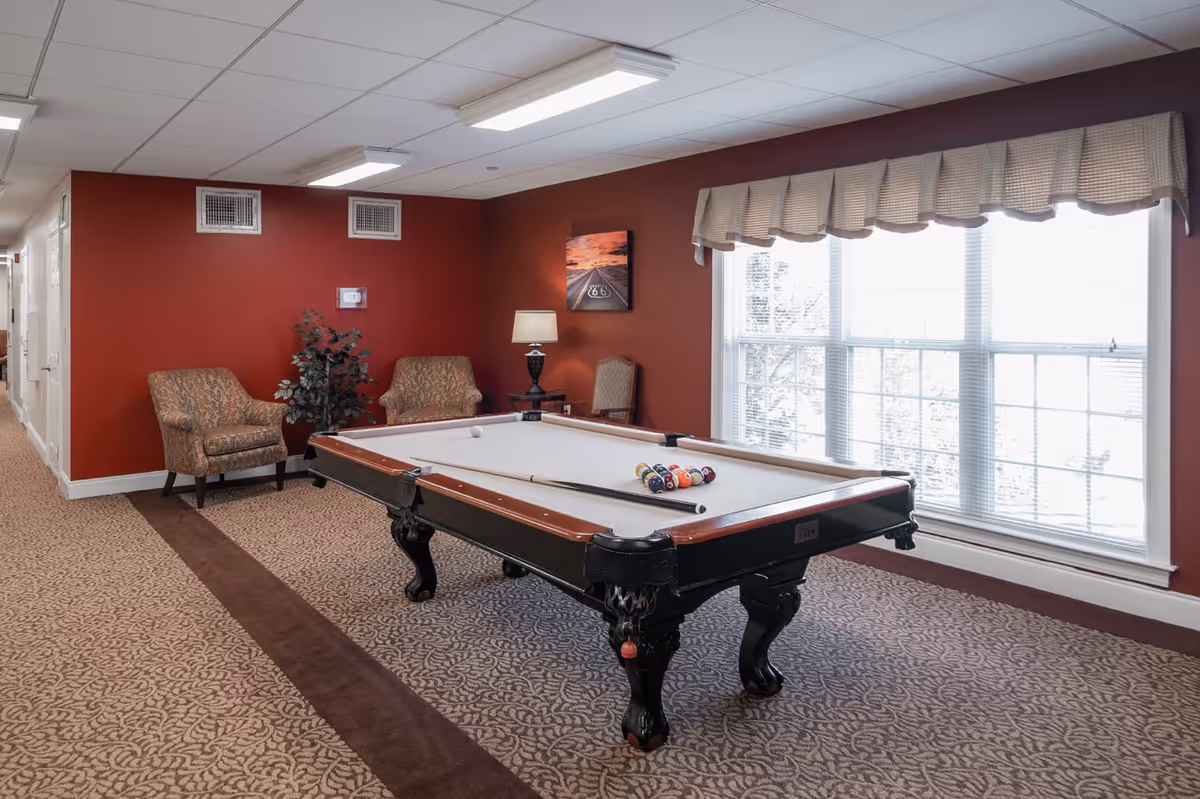A recreational room with a pool table set up for a game, two upholstered chairs, a floor lamp, a potted plant, and a large window with blinds and a valance letting in natural light. The walls are painted red and the floor is carpeted with a patterned design.