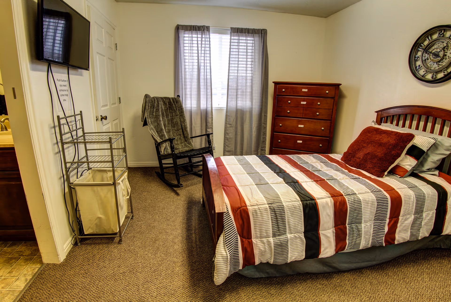 A bedroom with a bed covered in a striped comforter in shades of red, gray, black, and white. There is a large red pillow and several smaller pillows on the bed. A wooden dresser with multiple drawers is against the wall near a window with gray curtains. A black rocking chair with a gray blanket draped over it is positioned near the window. A flat-screen TV is mounted on the wall opposite the bed. The floor is carpeted, and part of a bathroom sink and mirror are visible through an open doorway.