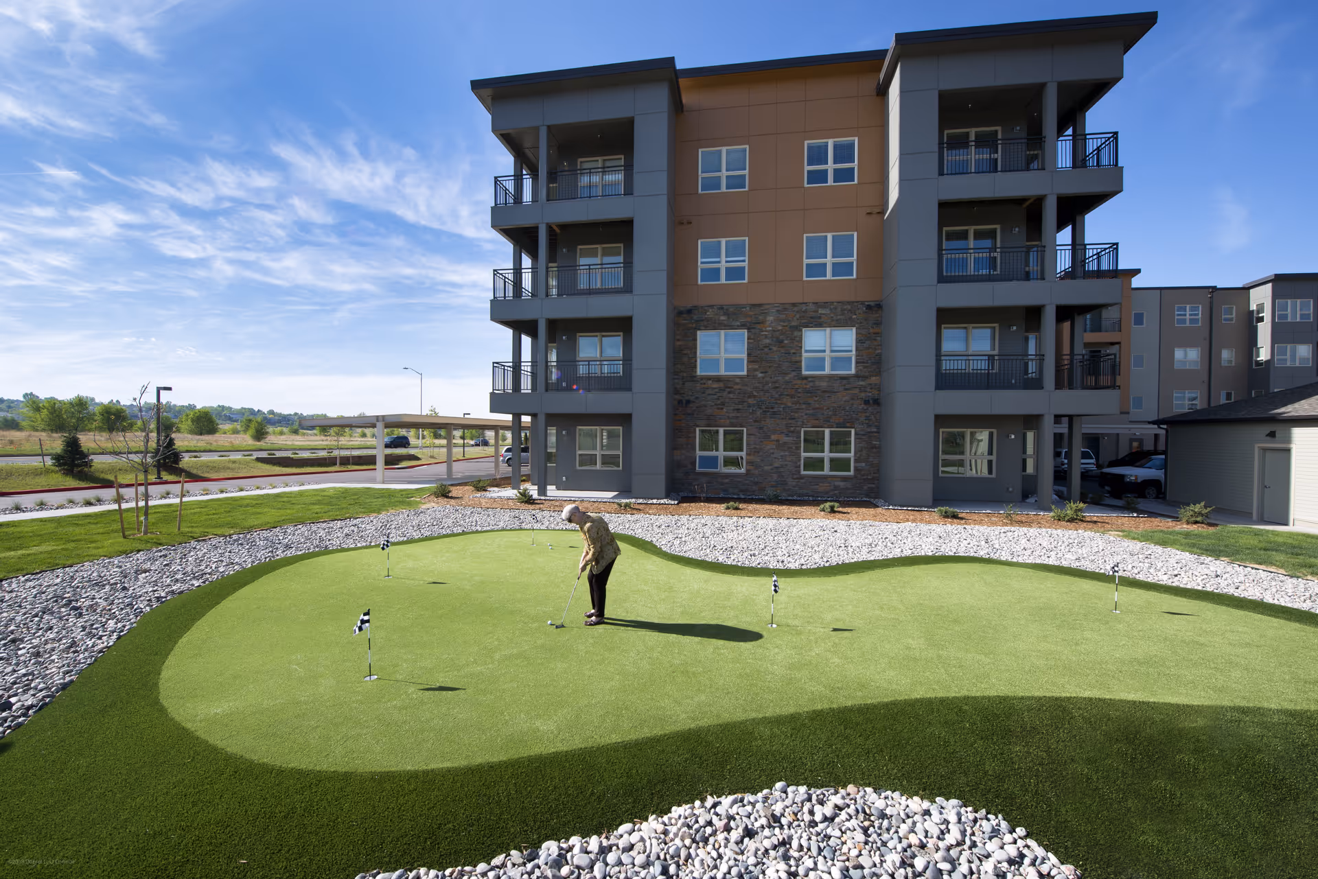 An elderly person playing golf on a putting green outside a modern multi-story residential building under a blue sky with scattered clouds.