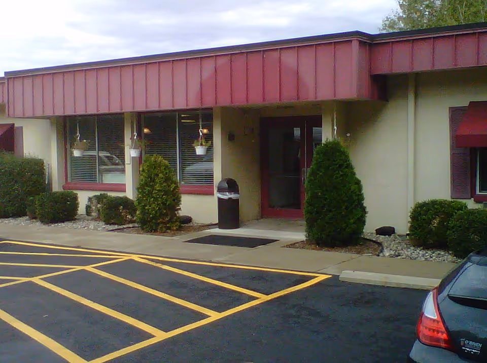 Exterior view of a single-story building entrance with a maroon awning and door frame, surrounded by small bushes and plants. There is a parking lot with yellow-striped parking spaces in front, and a black car is partially visible on the right side.
