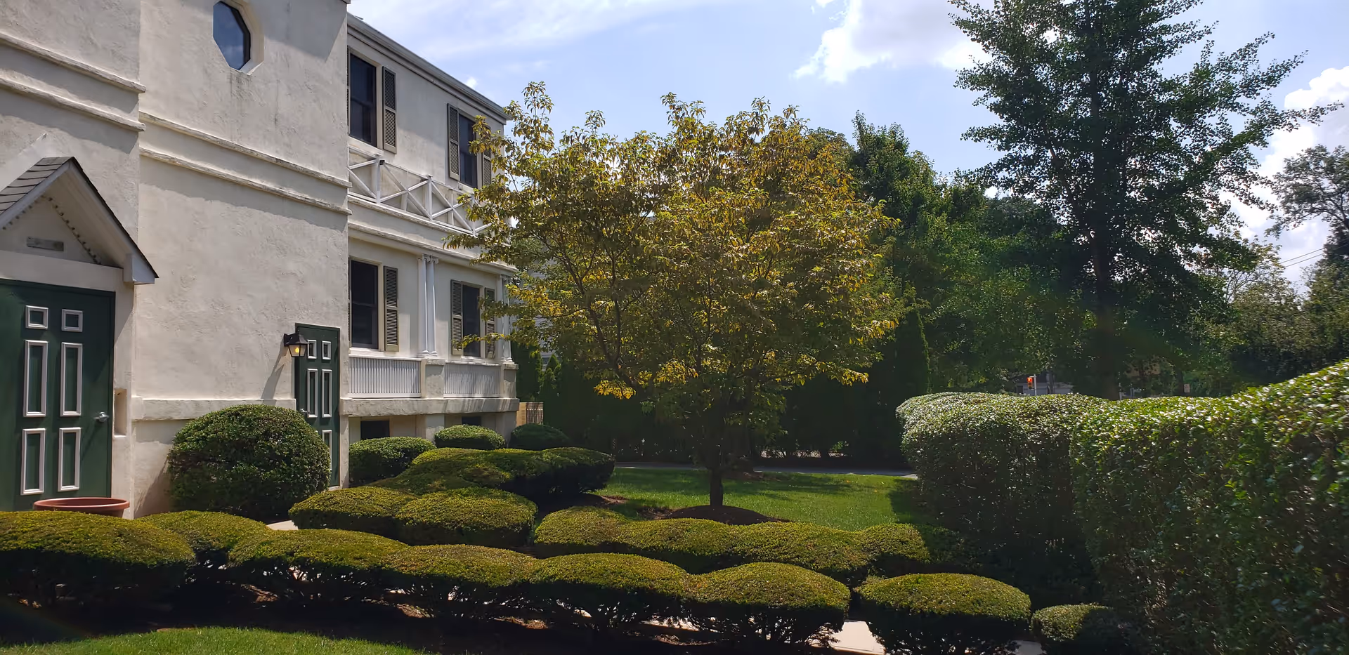 Exterior view of a senior living facility showing a white building with green doors, neatly trimmed bushes, a tree, and a well-maintained lawn under a partly cloudy sky.
