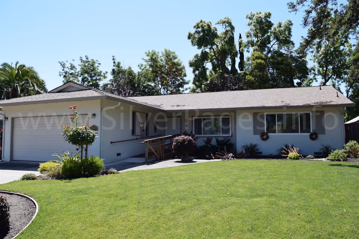 Single-story residential building with a light-colored exterior, a garage on the left, a wheelchair ramp leading to the front door, and a well-maintained green lawn with various plants and small bushes in front. Trees are visible in the background under a clear blue sky.