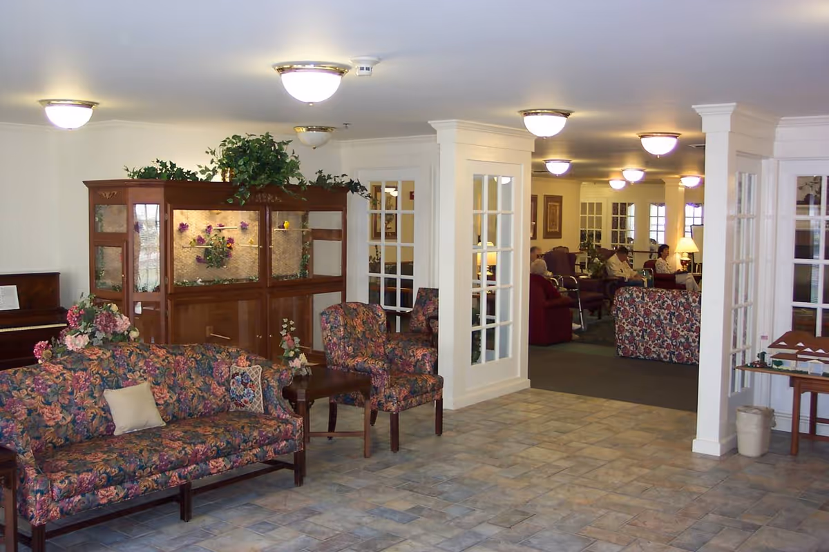 A spacious senior living facility common area with floral patterned sofas and chairs, a wooden display cabinet with decorative plants and flowers, a piano in the corner, and several seniors seated in the background. The room has tiled flooring, white walls, and ceiling lights.