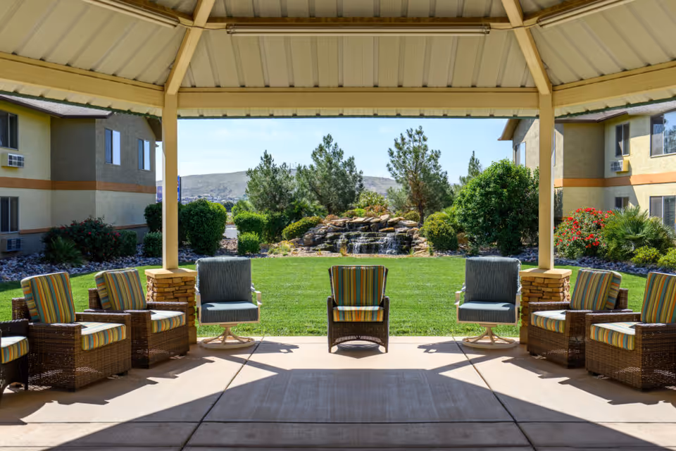 Covered patio with wicker and cushioned chairs facing a grassy courtyard and a small rock waterfall between two apartment buildings.