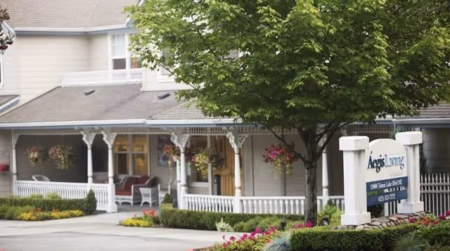 Exterior view of Aegis Living Kirkland facility showing a porch with white railings and hanging flower baskets, a tree in the foreground, and a sign with the facility name and contact information.