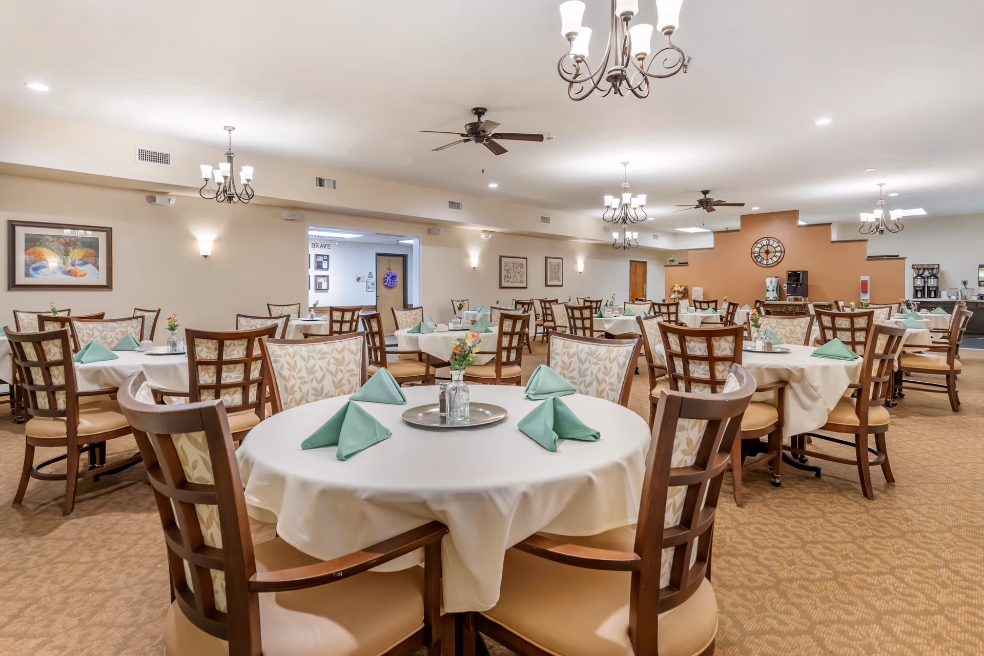 A spacious dining room with multiple round tables covered with white tablecloths and set with green folded napkins. Each table has a small centerpiece with flowers. The room is well-lit with chandeliers and ceiling fans, and there are framed artworks on the walls. In the background, there is a coffee and beverage station.