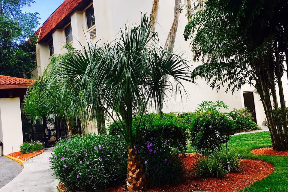 Outdoor garden area at Langdon Hall with palm trees, green shrubs, purple flowers, and a building with a red-tiled roof in the background under a blue sky.