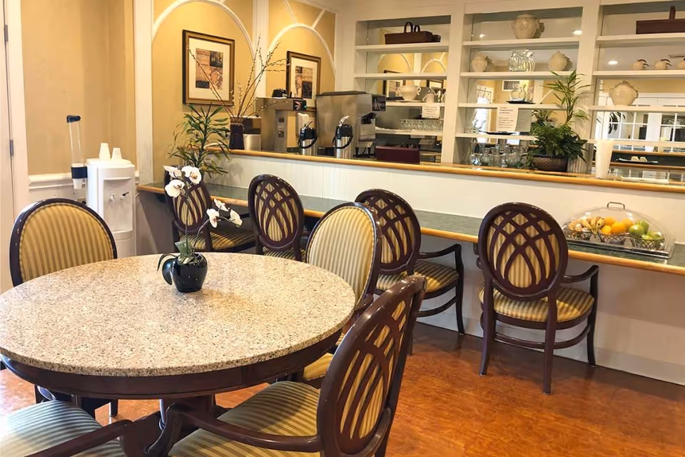 Round dining table with an orchid centerpiece, upholstered chairs, and a serving counter with a coffee station and fruit display in a dining area.