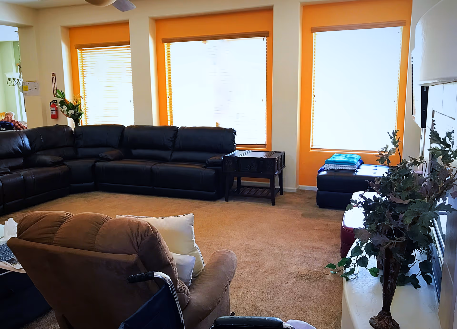 A living room in an assisted living home with a large black leather sectional sofa, a brown recliner chair, a small wooden side table, and a black ottoman with folded blankets on top. The room has three large windows with orange trim and blinds, beige carpet, and a vase with green leafy plants on a white surface.