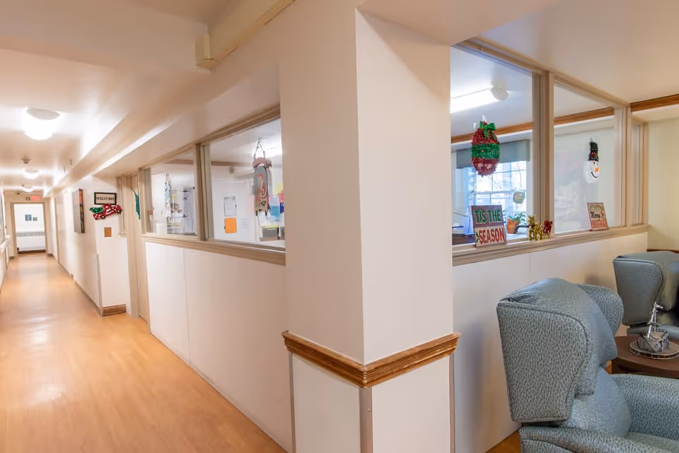 A well-lit hallway in a senior living facility with light wood flooring and white walls. On the right side, there is a seating area with blue upholstered chairs and a small table. The wall has large windows decorated with holiday-themed ornaments and signs, including one that says 'Tis the Season'. The hallway extends into the distance with doors and a 'Welcome' sign visible on the left side.