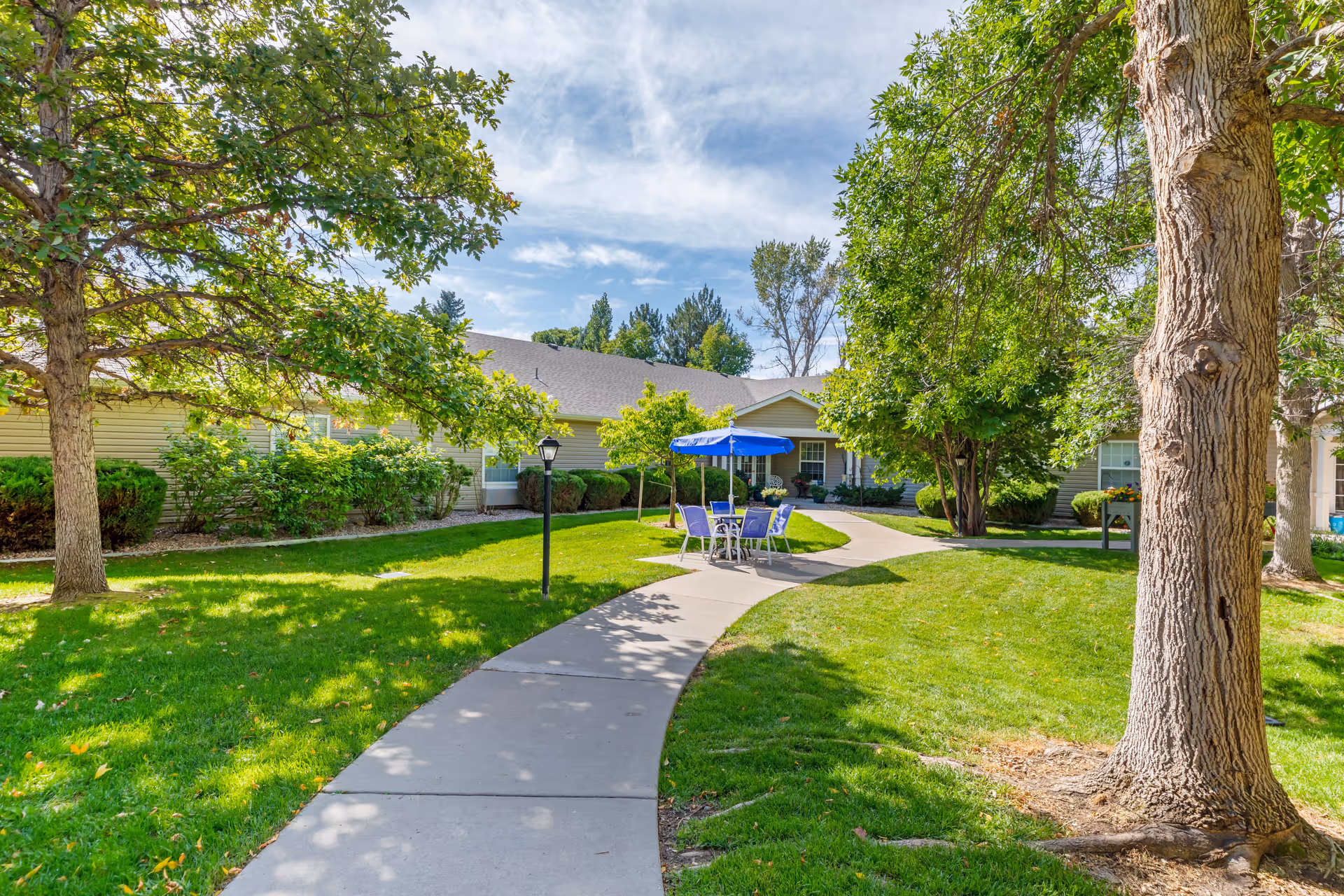 A sunny outdoor courtyard area at Brookdale North Loveland featuring a winding concrete pathway, green grass, several trees, and a small seating area with a table, chairs, and a blue umbrella.