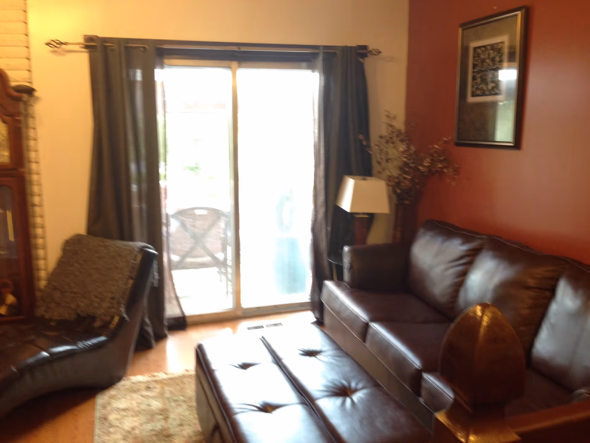 Cozy living room with a brown leather sofa and ottoman, chaise chair, lamp, and sliding glass doors with curtains.
