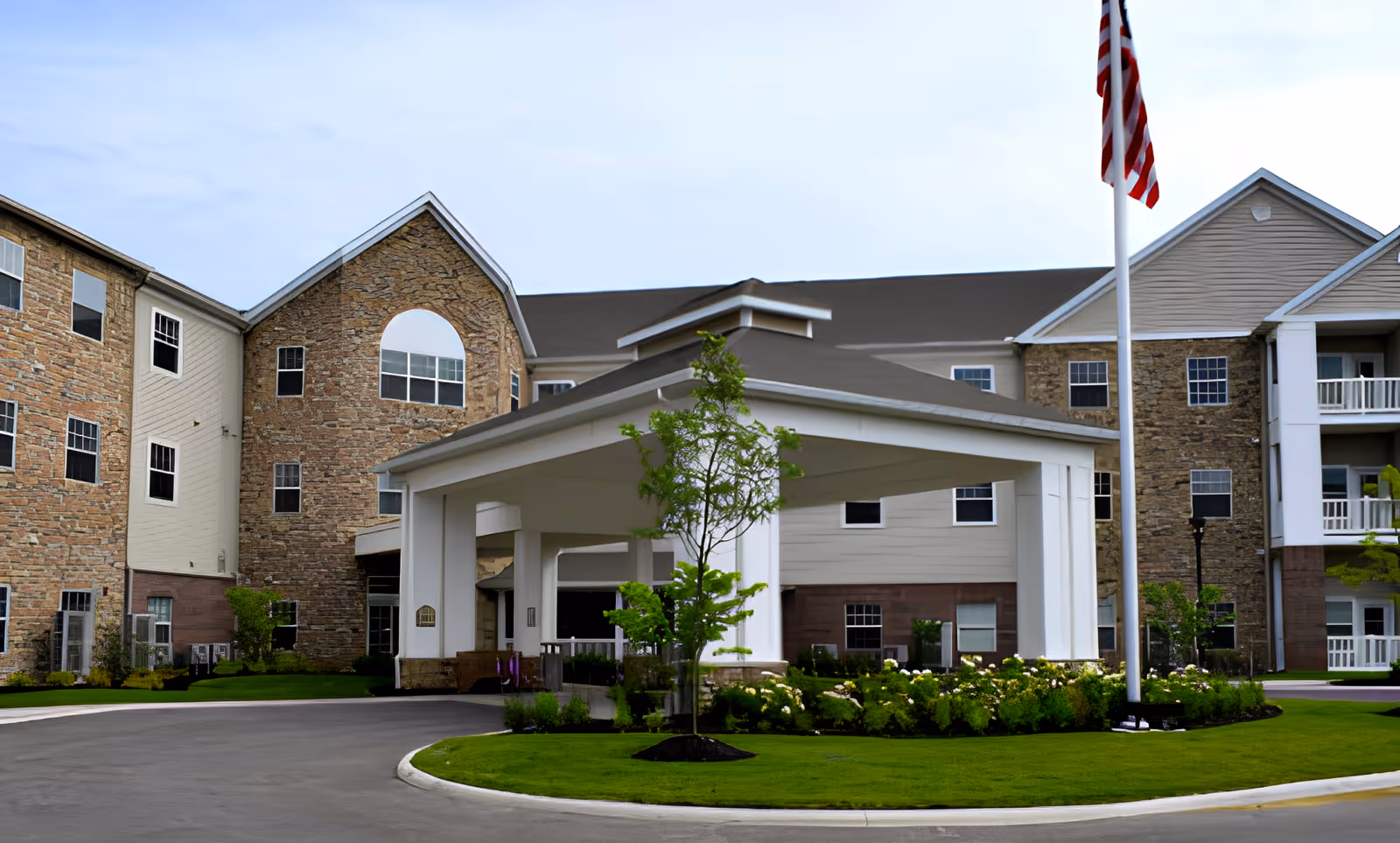 Front exterior view of Danbury Hudson facility showing a large building with stone and siding facade, a covered entrance with white pillars, a curved driveway, landscaped greenery, and an American flag on a flagpole.