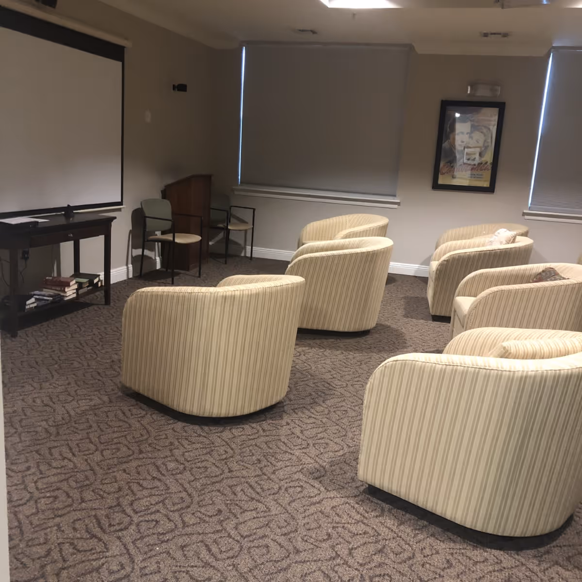 A small theater or media room with six beige striped armchairs arranged in two rows facing a large white projection screen. There are two additional chairs and a small podium in the corner near the screen. The room has carpeted flooring, two windows with closed blinds, and a framed movie poster on the wall.