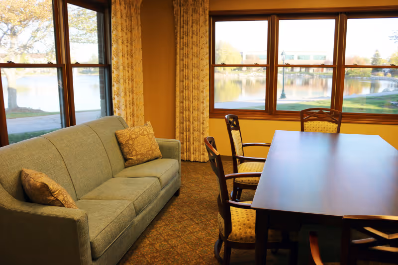 A cozy room with a green upholstered sofa with two patterned cushions, a wooden table surrounded by four wooden chairs with yellow patterned cushions, large windows with floral curtains showing a view of a lake and trees outside.