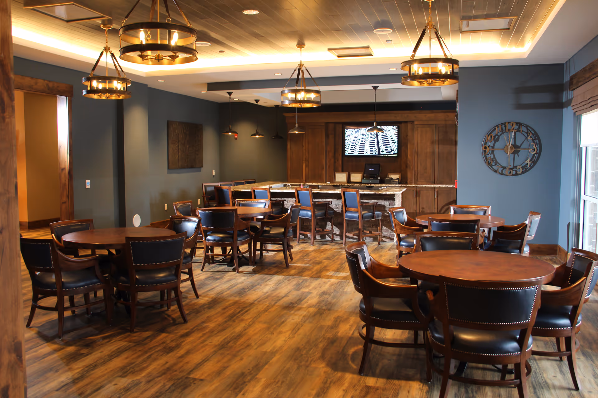 A spacious dining area in a senior living community with several round wooden tables surrounded by dark leather chairs. The room features wood flooring, blue-gray walls, and large windows with blinds. There is a counter with bar stools and a mounted TV on the back wall, along with decorative hanging lights and a large wall clock.