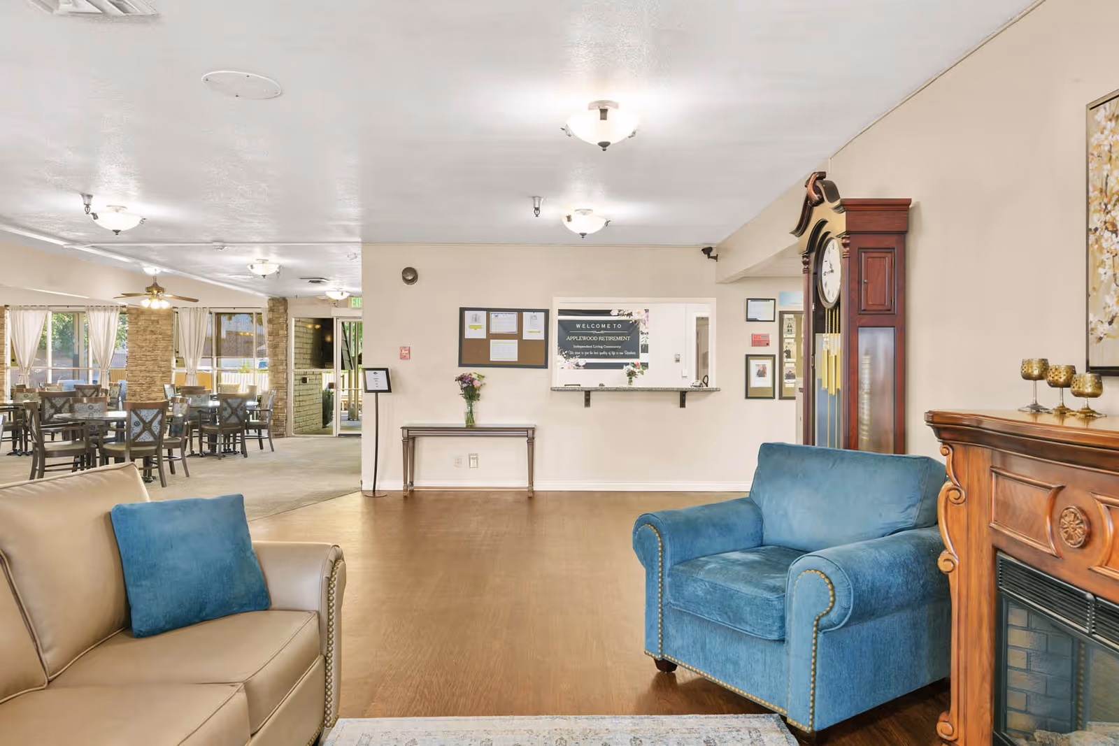 Interior view of a senior living facility lounge area with a beige sofa and a blue armchair in the foreground. There is a wooden grandfather clock and a fireplace with decorative items on the right. In the background, there is a reception desk with a welcome sign and a dining area with tables and chairs near large windows with curtains.