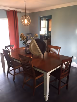 A dining room with a wooden table and six matching chairs. The table has decorative items including a globe and a vase with flowers. The room has blue walls, dark wood flooring, a hanging light fixture above the table, and red curtains on a window to the left. A kitchen area is visible through an open wall space.