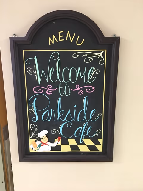 A decorative chalkboard sign with the word 'MENU' at the top and colorful cursive writing that says 'Welcome to Parkside Cafe'. The sign has a black frame and features a small illustration of a chef holding a loaf of bread and a drink at the bottom left corner.