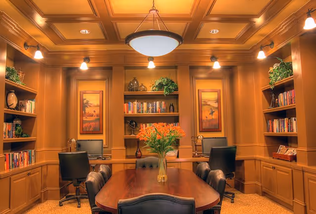 A cozy, wood-paneled room with a large wooden table surrounded by black leather chairs. The walls have built-in bookshelves filled with books and decorative items, and two framed paintings hang on the back wall. There are two computer workstations with black office chairs on either side of the room. A vase with orange flowers is placed in the center of the table, and warm lighting fixtures illuminate the space.