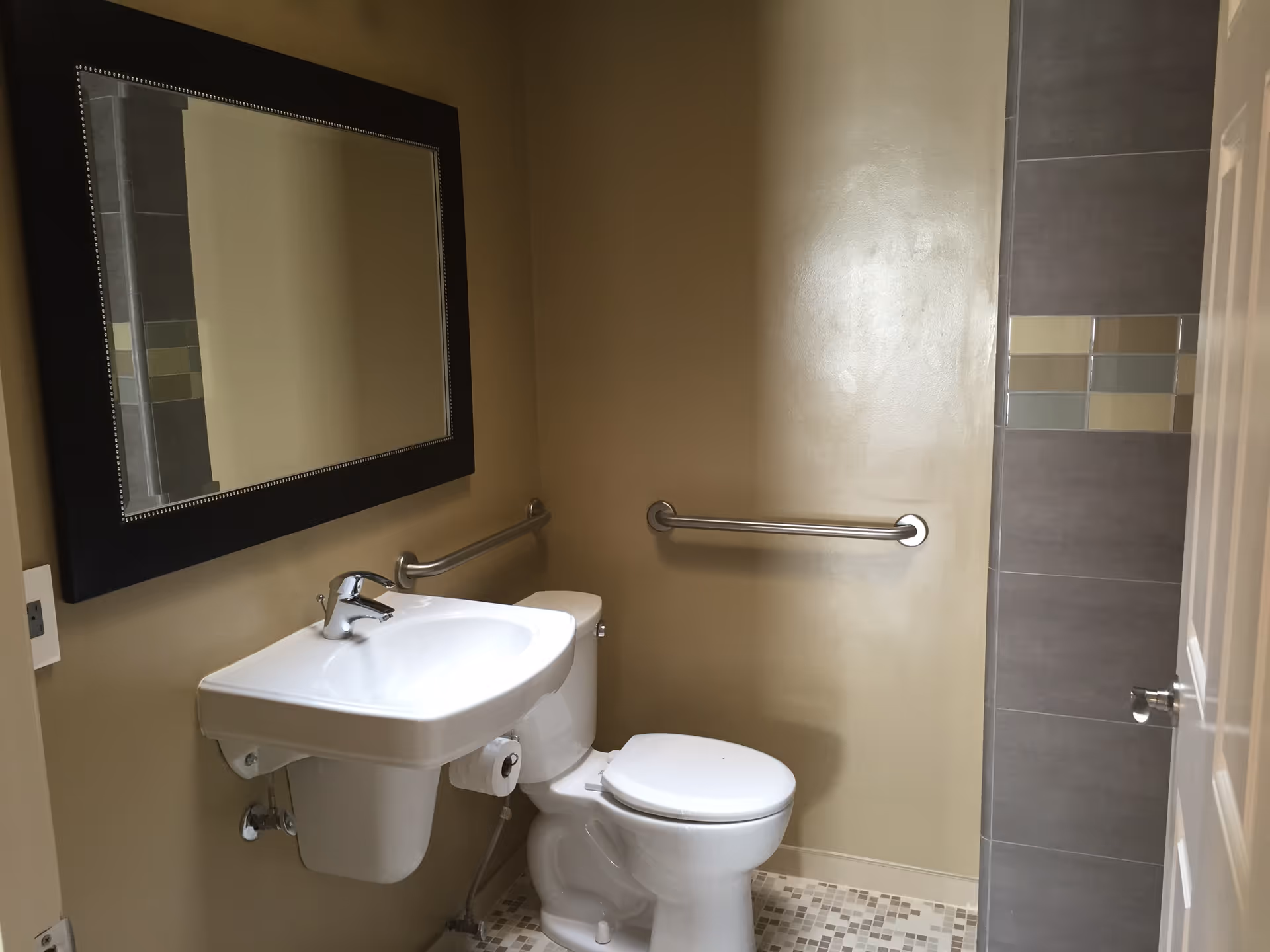 A small bathroom with a white wall-mounted sink, a toilet, a large rectangular mirror with a black frame above the sink, beige walls, a tiled floor with a mosaic pattern, and a grab bar on the wall next to the toilet. The edge of a tiled shower area with decorative tiles is visible on the right side.