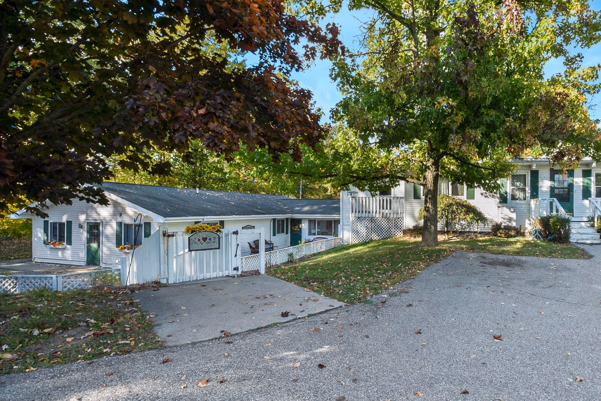 Exterior view of Caremore of Oxford facility showing a white building with green doors and window shutters, surrounded by trees with green and brown leaves. There is a white fence with a sign and a paved driveway in front.