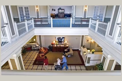Two-story senior living common area with seating, a reception desk, and people including one person in a wheelchair viewed from an upper balcony.