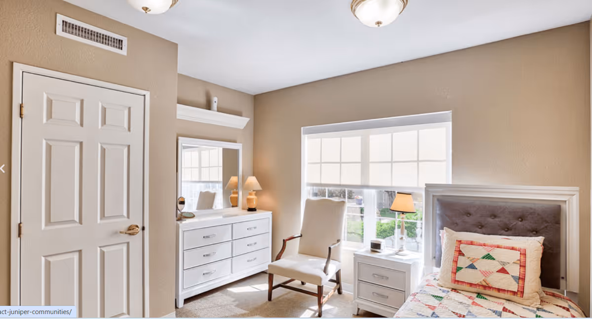 A bright bedroom with beige walls featuring a white dresser with a mirror and two lamps, a white armchair, a window with white blinds, a nightstand with a lamp and clock, and a bed with a colorful patchwork pillow and gray tufted headboard.