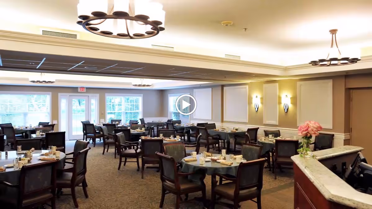 A spacious dining room with multiple round tables covered with dark tablecloths, each set with cups, glasses, and napkins. The room has large windows letting in natural light, beige walls with decorative panels, and ceiling lights with circular fixtures. A reception desk with a vase of pink flowers is visible in the foreground.