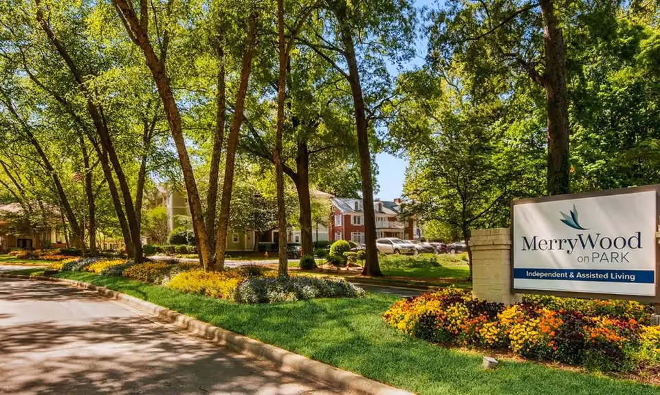 Outdoor view of MerryWood on Park senior living facility with a landscaped garden, trees, and a sign that reads 'MerryWood on Park Independent & Assisted Living'. Residential buildings and parked cars are visible in the background.