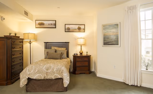 Well-lit furnished bedroom with a single bed, nightstand, dresser, lamps, and framed artwork.