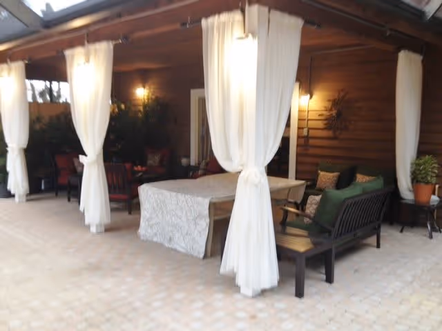Covered outdoor patio area with white curtains tied to wooden posts, a table with a patterned tablecloth, cushioned benches, chairs, and potted plants against a wooden wall.