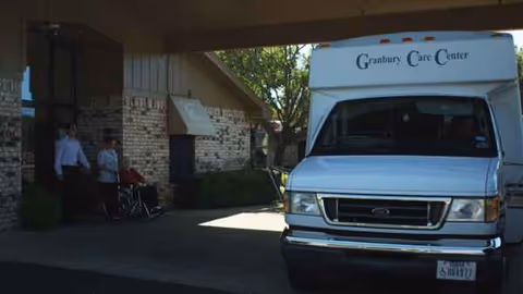 A white Granbury Care Center vehicle parked at the building entrance with a few people nearby.