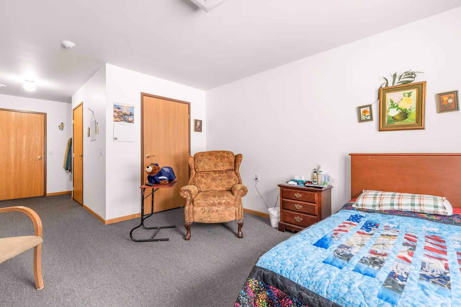 A senior living facility bedroom with a single bed covered in a colorful quilt, a wooden headboard, and a plaid pillow. Next to the bed is a wooden nightstand with various personal items on top. An upholstered armchair with a floral pattern sits near the center of the room, next to a small table holding a stuffed bear. The room has light-colored walls adorned with framed floral paintings and a calendar. There are two wooden doors visible, and the floor is carpeted in gray.