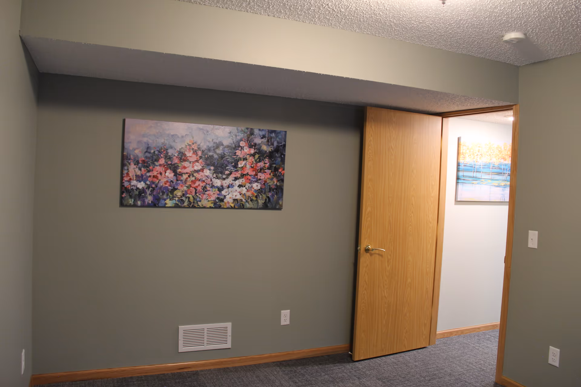 An interior room with grayish-green walls and a carpeted floor. A wooden door is partially open, revealing a hallway with a light blue wall and a painting of trees and water. On the main room's wall, there is a colorful painting of flowers. The room has a vent and electrical outlets near the baseboard.