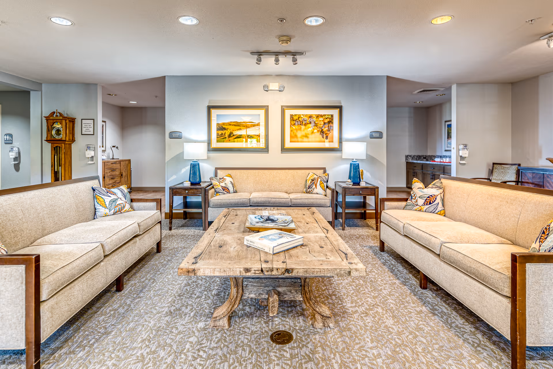 A well-lit senior living facility common area with three beige sofas arranged around a rustic wooden coffee table. The sofas have decorative pillows with leaf patterns. Two blue table lamps sit on wooden side tables flanking the middle sofa. Two framed landscape pictures hang on the wall above the middle sofa. A grandfather clock is visible on the left side near a hallway entrance.