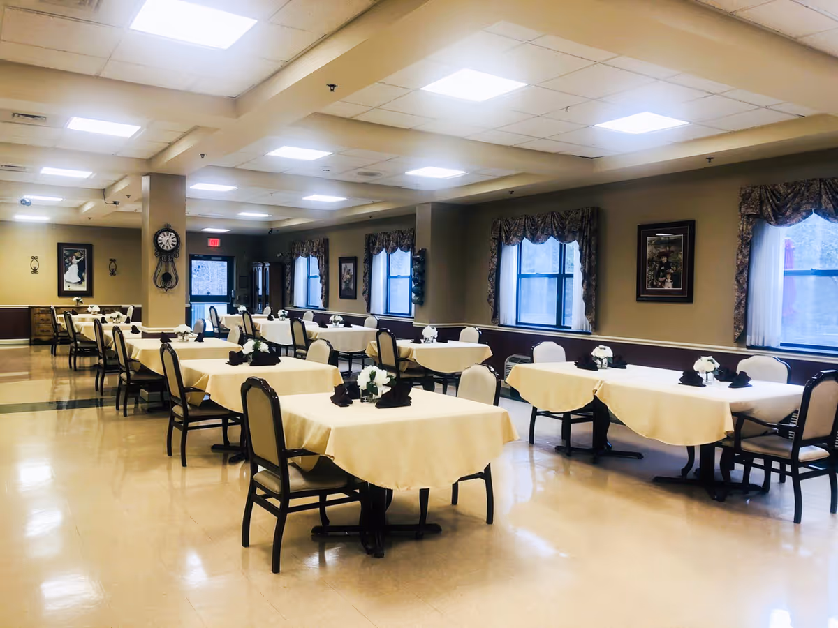 A spacious dining room with multiple tables covered in beige tablecloths, each set with white floral centerpieces and dark folded napkins. The room has several windows with patterned valances, framed artwork on the walls, and a clock mounted on a pillar. The floor is polished and the ceiling has recessed lighting.