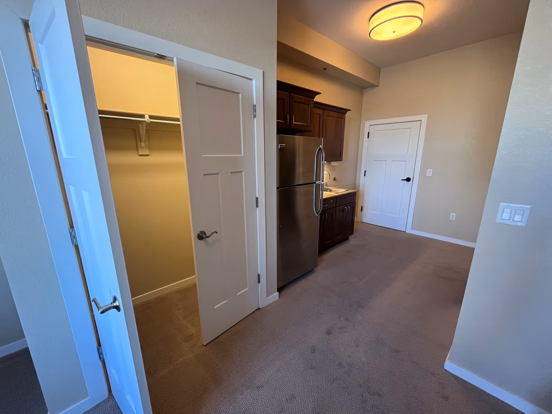 Interior view of a small kitchen area with a stainless steel refrigerator, dark wooden cabinets, and a sink. To the left, there is an open door revealing a walk-in closet with a hanging rod and shelf. The room has beige walls, carpeted floor, and a ceiling light fixture.