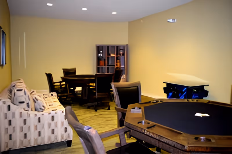 Interior room with a patterned sofa on the left, a poker table with chairs in the foreground, and a round table with chairs in the background against a beige wall.