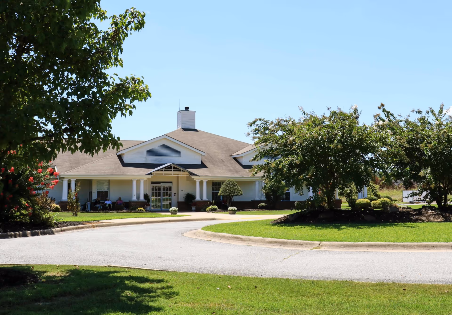 Front exterior view of a single-story senior living facility building with a covered entrance, white columns, and a chimney. The building is surrounded by well-maintained green lawns, trees, and shrubs under a clear blue sky. There are a few people sitting on benches near the entrance.