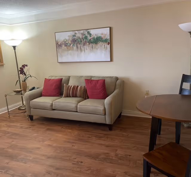 Small living room with a beige sofa topped with red pillows, wall art above, floor lamps, a side table with a plant, and a round dining table on wood flooring.
