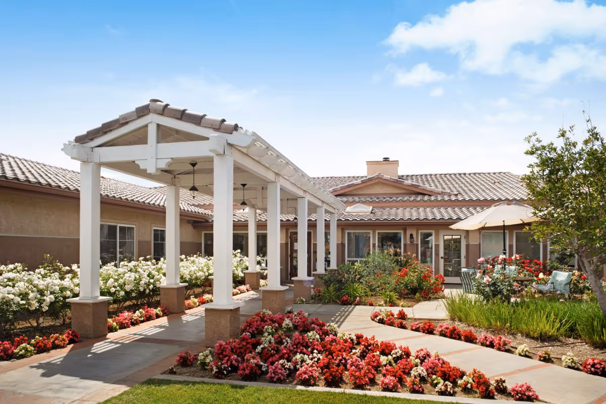Outdoor garden area at a senior living facility with a covered walkway supported by white pillars, surrounded by colorful flower beds and greenery. The building has a tiled roof and beige walls, with patio seating and an umbrella visible on the right side under a clear blue sky.