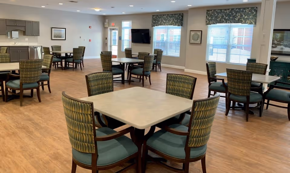 A spacious dining room with multiple square tables and green cushioned chairs arranged neatly. The room has large windows with patterned valances, a wall-mounted TV, and light wood flooring. There is a small kitchenette area with cabinets and a sink in the background.