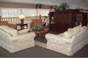 A cozy living room area with two cream-colored sofas facing each other around a round wooden coffee table with a plant centerpiece. There is a wooden entertainment center with a television and shelves, a small side table with a lamp, and large windows with blinds letting in natural light.
