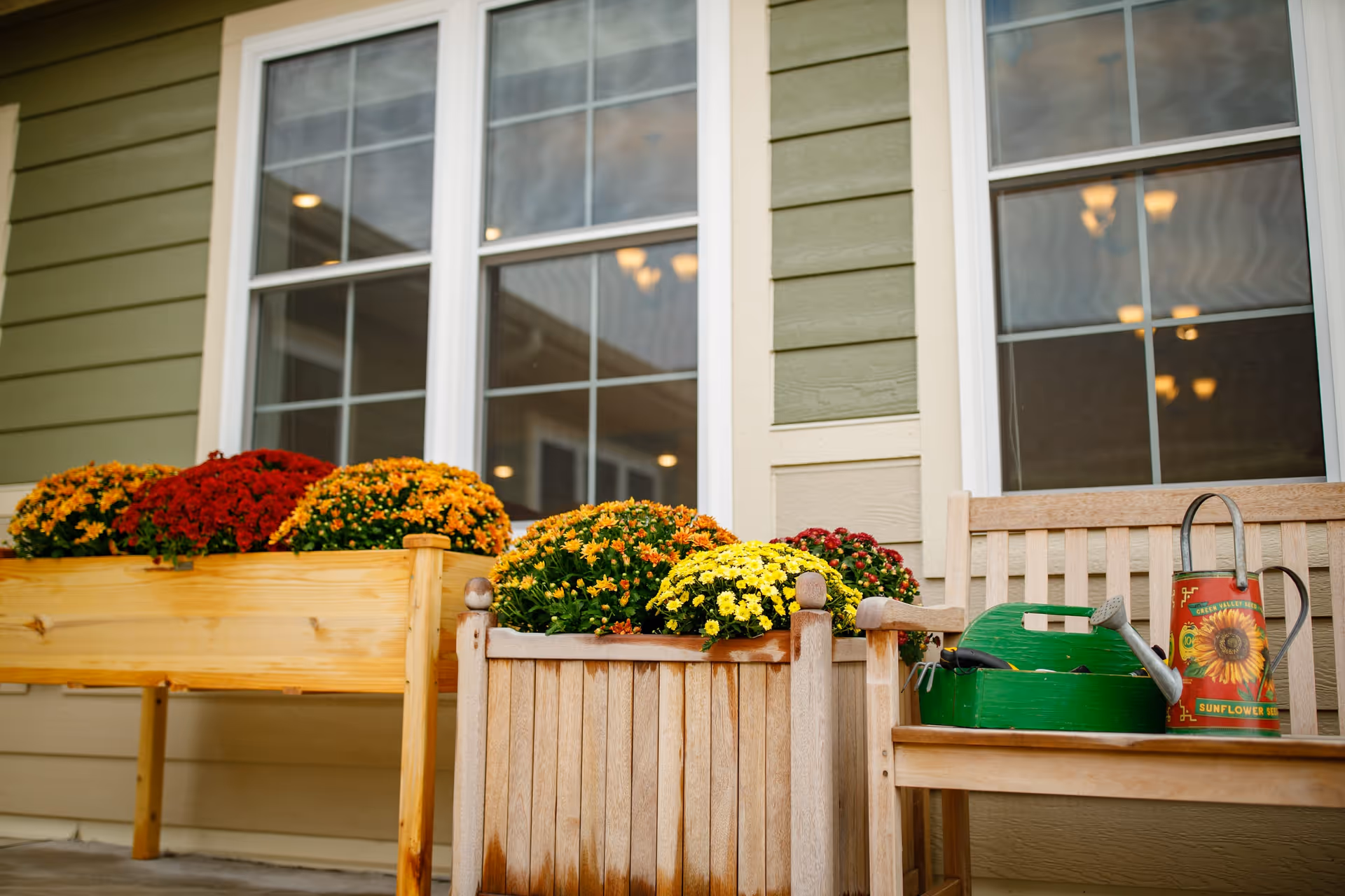A wooden planter box and a wooden bench with colorful flowers including red, orange, yellow, and green foliage placed outside a building with green siding and white-framed windows. On the bench, there is a green gardening tool caddy and a red watering can with sunflower seed branding.