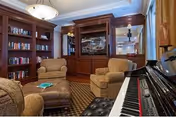 Cozy living room area with built-in wooden bookshelves filled with books, two upholstered armchairs, an ottoman, and a piano keyboard in the foreground. The room features warm lighting and a carpeted floor with a patterned rug.