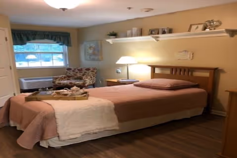 A cozy bedroom with a single bed covered in a pinkish-brown bedspread and a white throw blanket. There is a wooden headboard and a pillow on the bed. A tray with a teapot and cups is placed on the bed. Next to the bed is a wooden nightstand with a lamp and a telephone. A patterned armchair is positioned near a window with green valance curtains. Above the bed, a white shelf holds decorative items and framed pictures. The room has wooden flooring and soft yellow walls.